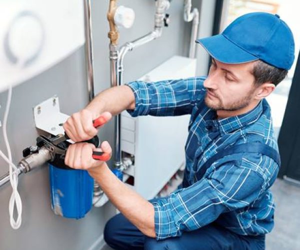 Young man in workwear using pliers while installing water filtration system in the kitchen of client